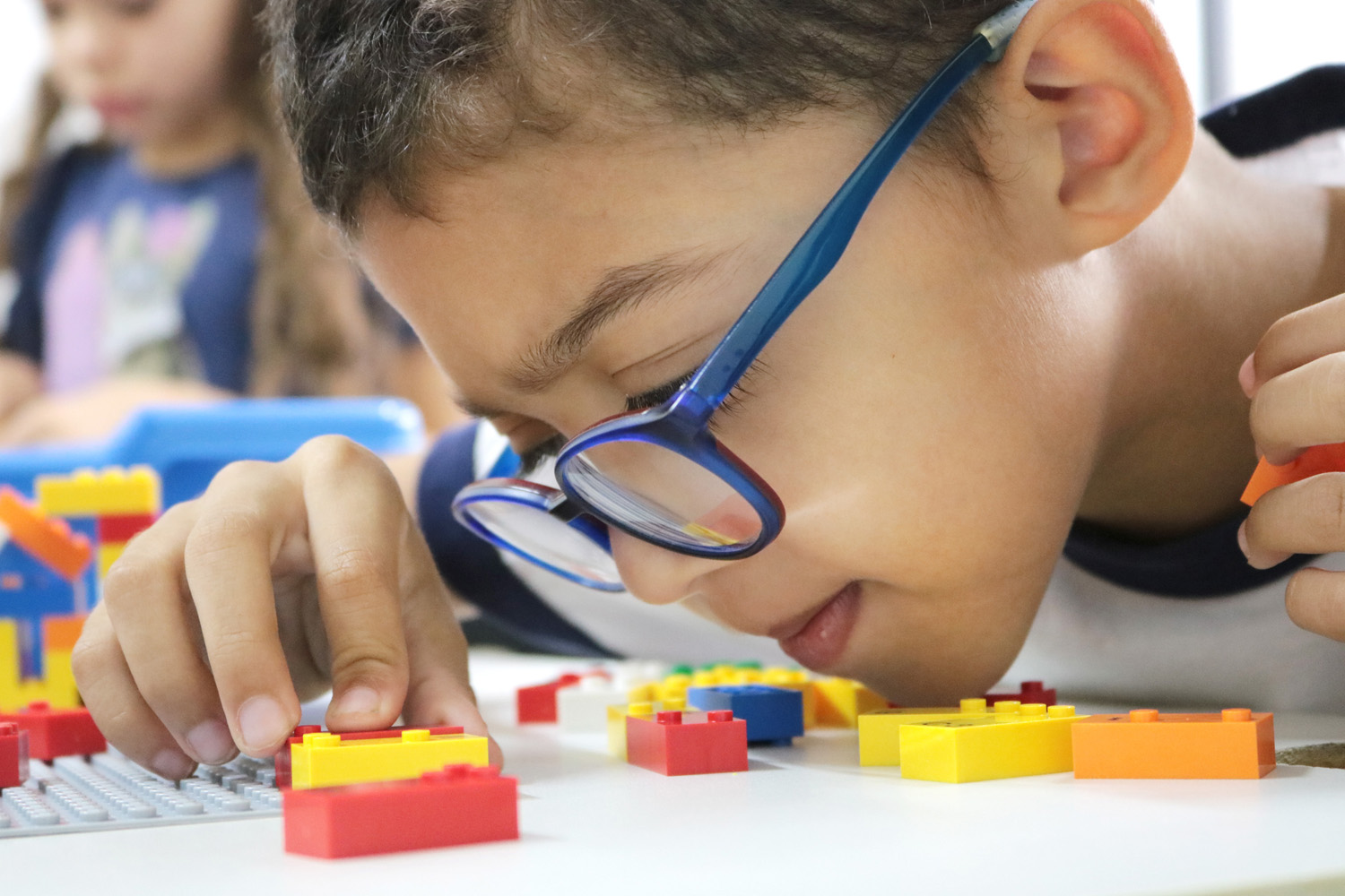 Descrição da imagem: foto de um garoto manuseando peças do LEGO Braille Bricks. Ele está de perfil, usa óculos e tem o rosto bem próximo às peças, com o queixo sobre a mesa.