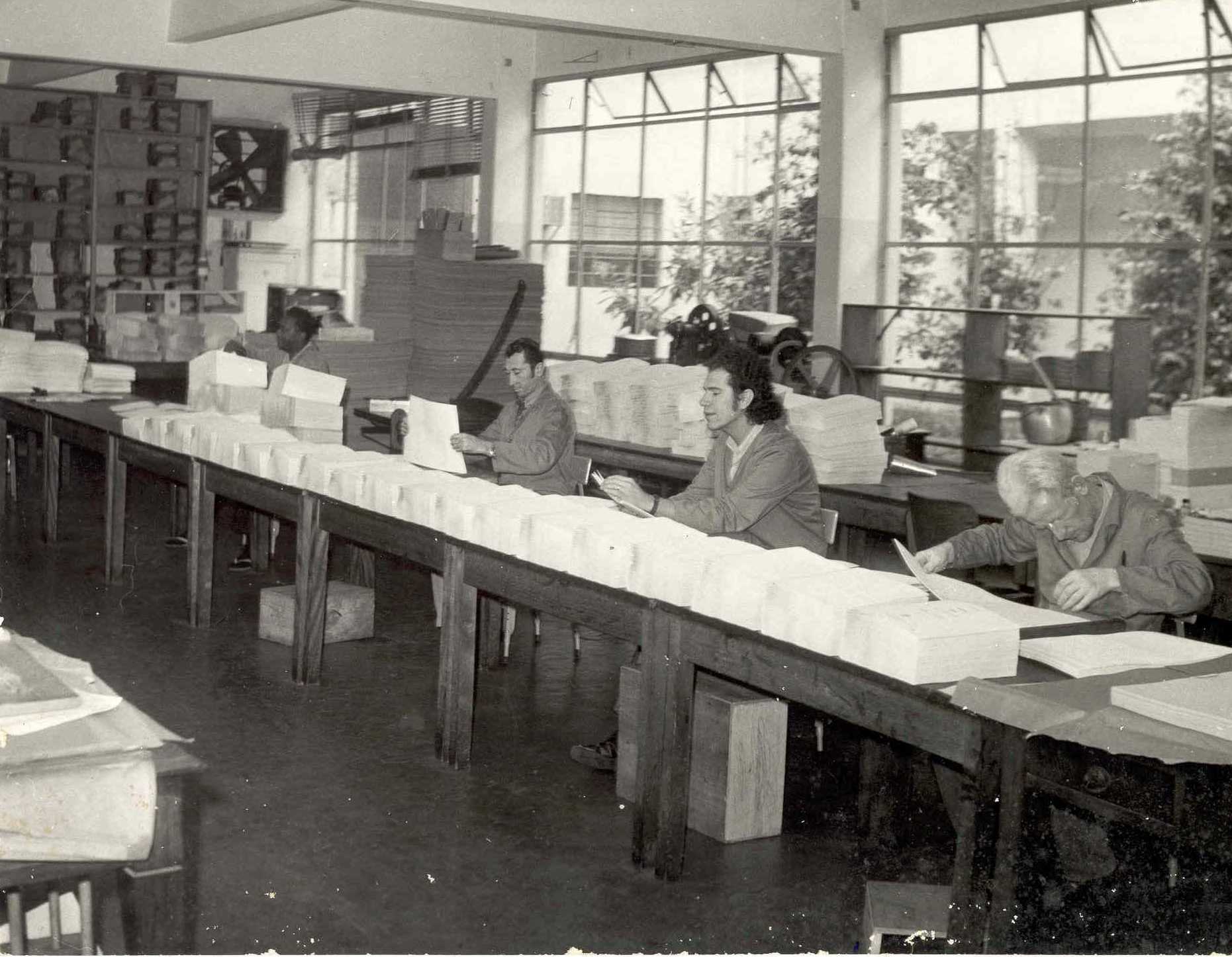 Descrição da imagem: foto em preto e branco de quatro homens manuseando pilhas de papel em branco sobre uma bancada.