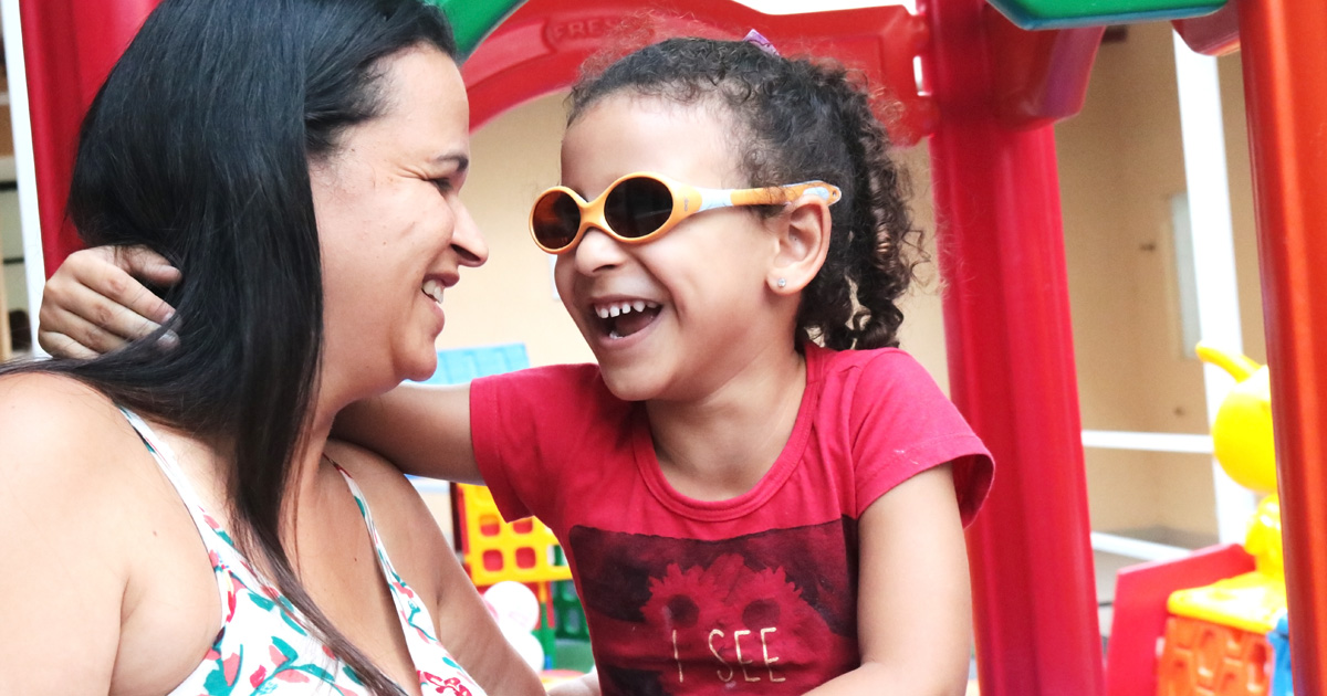 Descrição da imagem: foto de Julia com o braço sobre os ombros da mãe Alessandra no parquinho da Fundação Dorina. Elas se olham e sorriem.