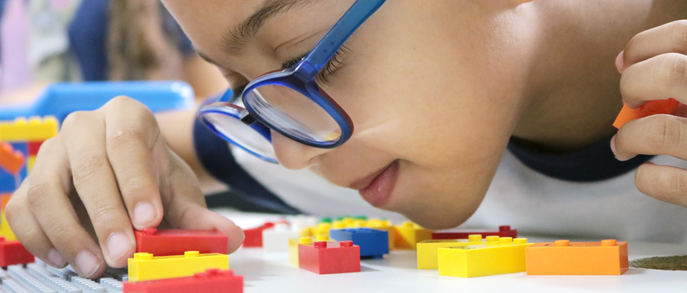 Descrição da imagem: foto de um menino de óculos manuseando pequenos blocos coloridos de montar. Ele tem o rosto bem próximo à mesa.