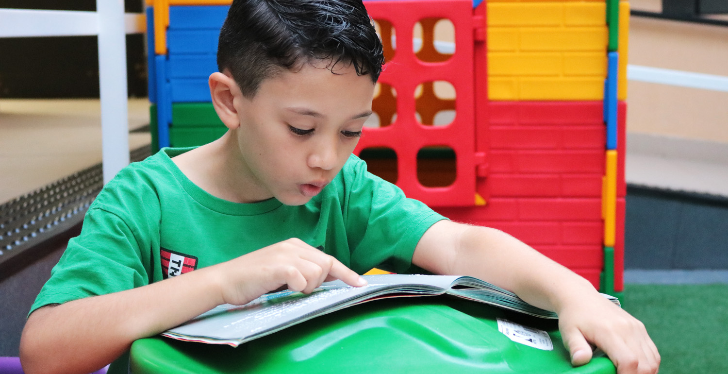 Descrição de imagem: foto de Gustavo lendo um livro aberto à sua frente. Ele tem o dedo indicador apontado para a página.