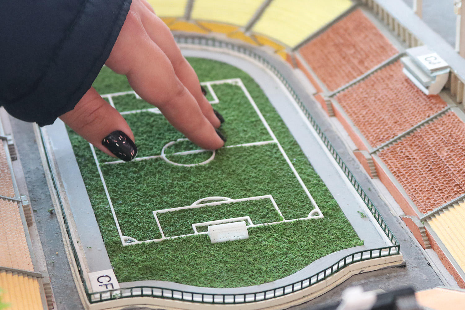 Descrição da imagem: foto da mão de Carla tocando a miniatura do campo do Pacaembu, de aproximadamente 10 cm. Na foto é possível ver também parte das arquibancadas do estádio em miniatura.