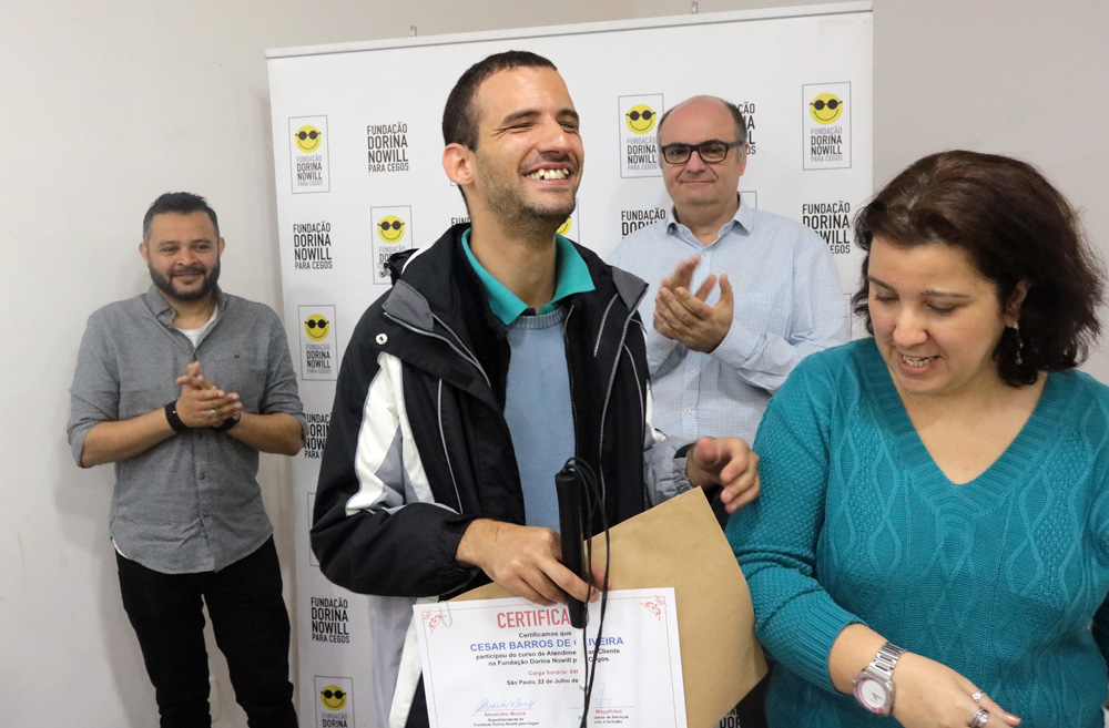 Descrição da imagem: foto de um aluno do curso de Atendimento ao Cliente sorrindo de olhos fechados. Ele segura uma bengala e o seu certificado com uma mão, e a outra está encostada no ombro de uma moça. Atrás deles, dois homens aplaudem.