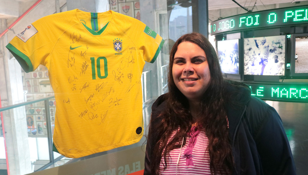 Descrição da imagem: foto de Carla posando ao lado da camisa 10 da jogadora Marta, autografada pelas companheiras de Seleção Brasileira. Carla olha pra frente e sorri.