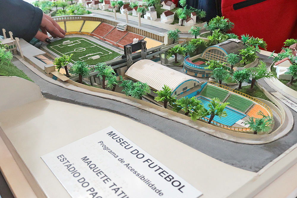Descrição da imagem: foto da maquete tátil do estádio do Pacaembu, incluindo o clube com piscina e quadras cobertas atrás do campo e as casas da vizinhança.