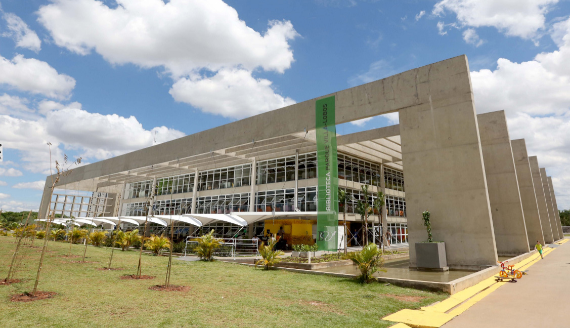 Descrição da imagem: foto do prédio da Biblioteca Parque Villa-Lobos em um dia de céu azul e nuvens brancas. O prédio tem largas colunas paraleleas no lado direito e um banner verde do teto ao chão com o título "Biblioteca Parque Villa-Lobos". Ao lado direito, um menino caminha em direção a uma pequena bicicleta.