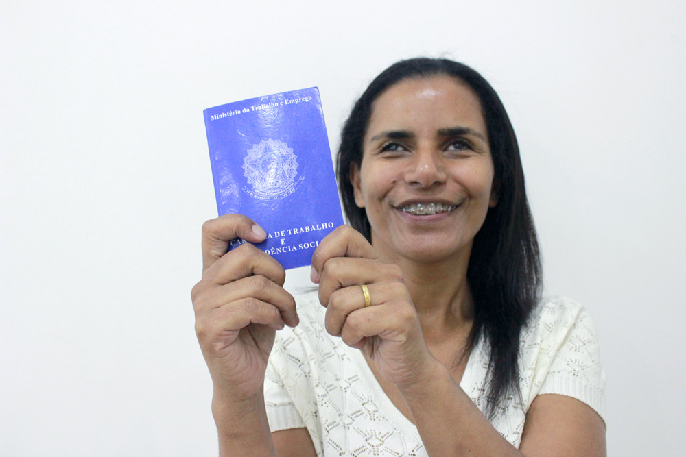 Descrição da imagem: foto de mulher segurando carteira de trabalho, apontando em direção à câmera. Ela olha pra frente e sorri.