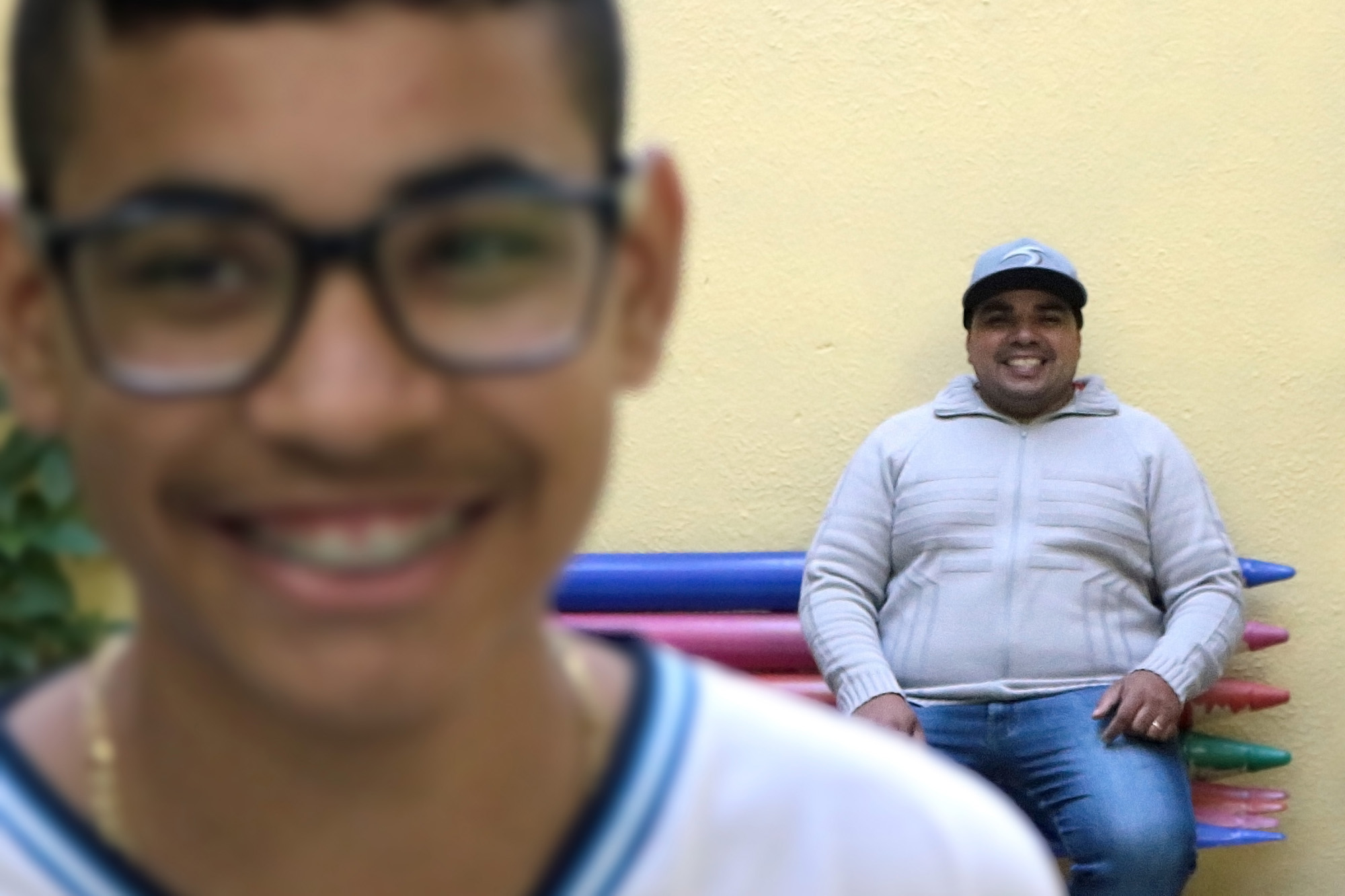 Descrição da imagem: foto de Iago e Moisés sorrindo. Iago está retratado do pescoço pra cima, levemente desfocado. Moisés está cerca de três metros atrás dele, sentado num banco colorido e olhando para o filho. O menino usa óculos e aparelho odontológico..