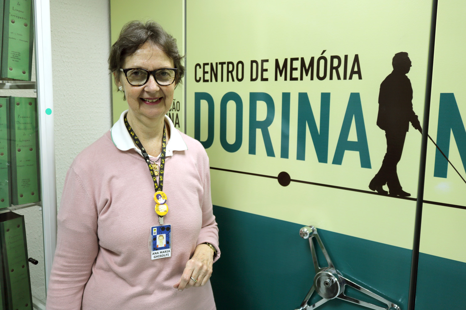 Descrição da imagem: Foto de Ana no Centro de memória da Fundação Dorina. Ela está olhando para foto e sorrindo. Ao fundo: Centro de Memória Dorina.
