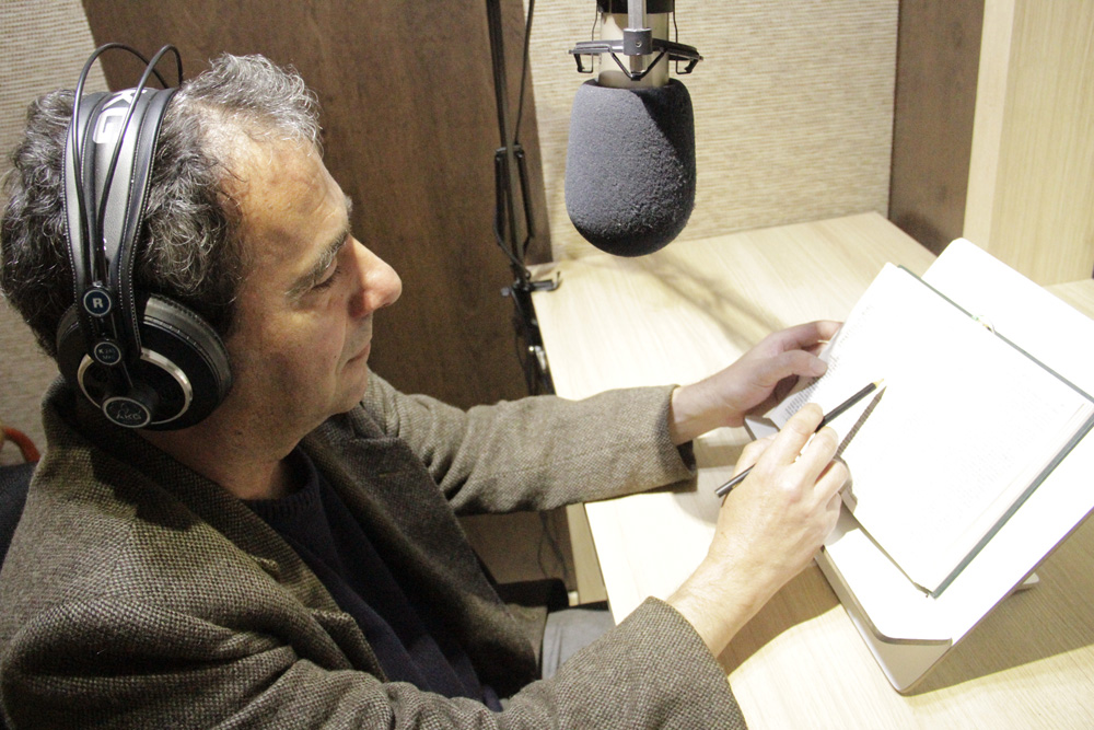 Descrição da imagem: foto de um homem de perfil com um livro aberto à sua frente. Ele toca a página com um lápis, usa fones de ouvido e tem um microfone de estúdio posicionado à sua frente.