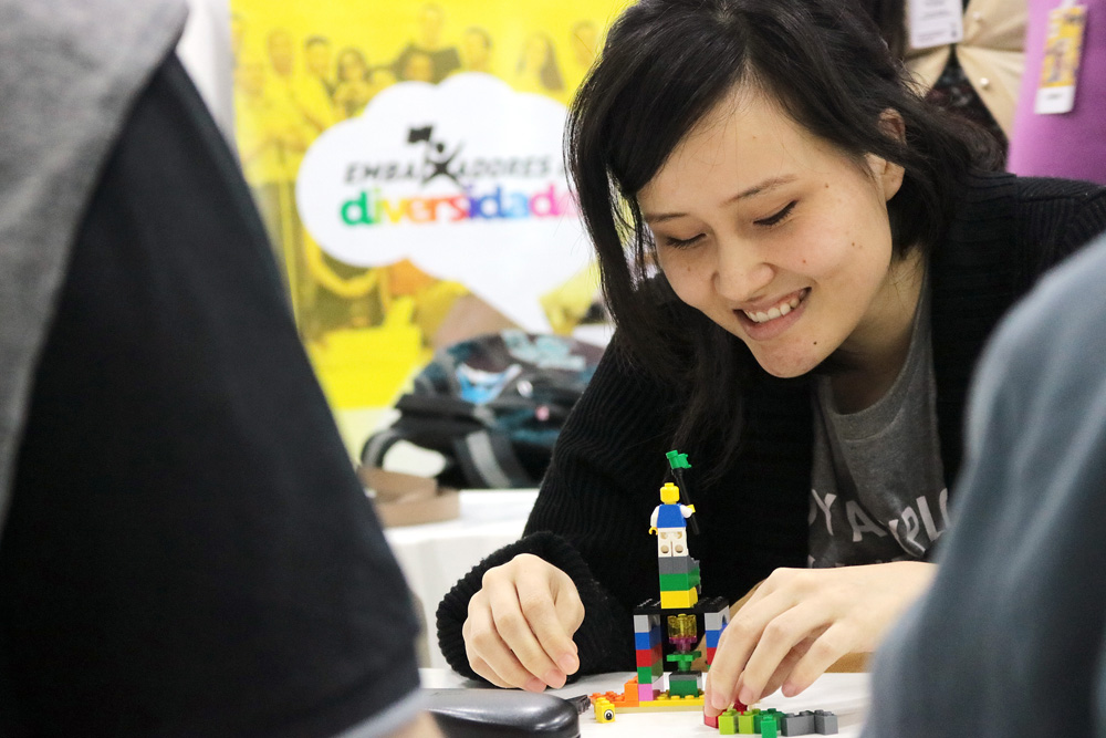 Descrição da imagem: foto de moça de traços orientais manuseando peças de Lego e sorrindo. Ao fundo há um banner do projeto "Embaixadores da Diversidade"