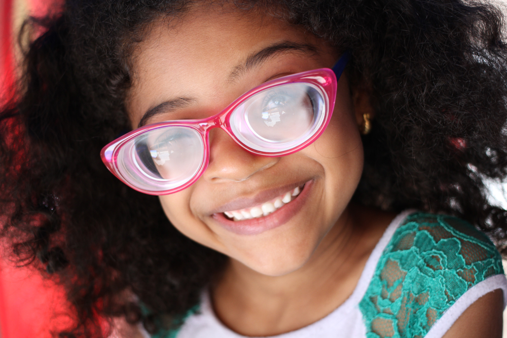 Descrição da imagem: foto de uma garotinha sorridente de óculos cor-de-rosa. Ela aparente ter cerca de 6 anos, é negra, tem cabelos cacheados na altura dos ombros e usa camiseta branca com mangas verdes.
