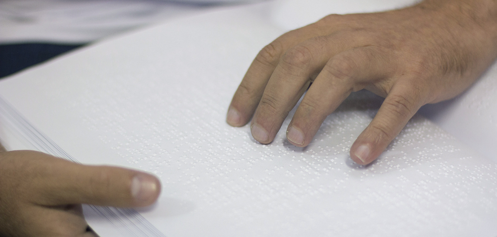 Descrição da imagem: foto de duas mãos tateando uma folha em braille.