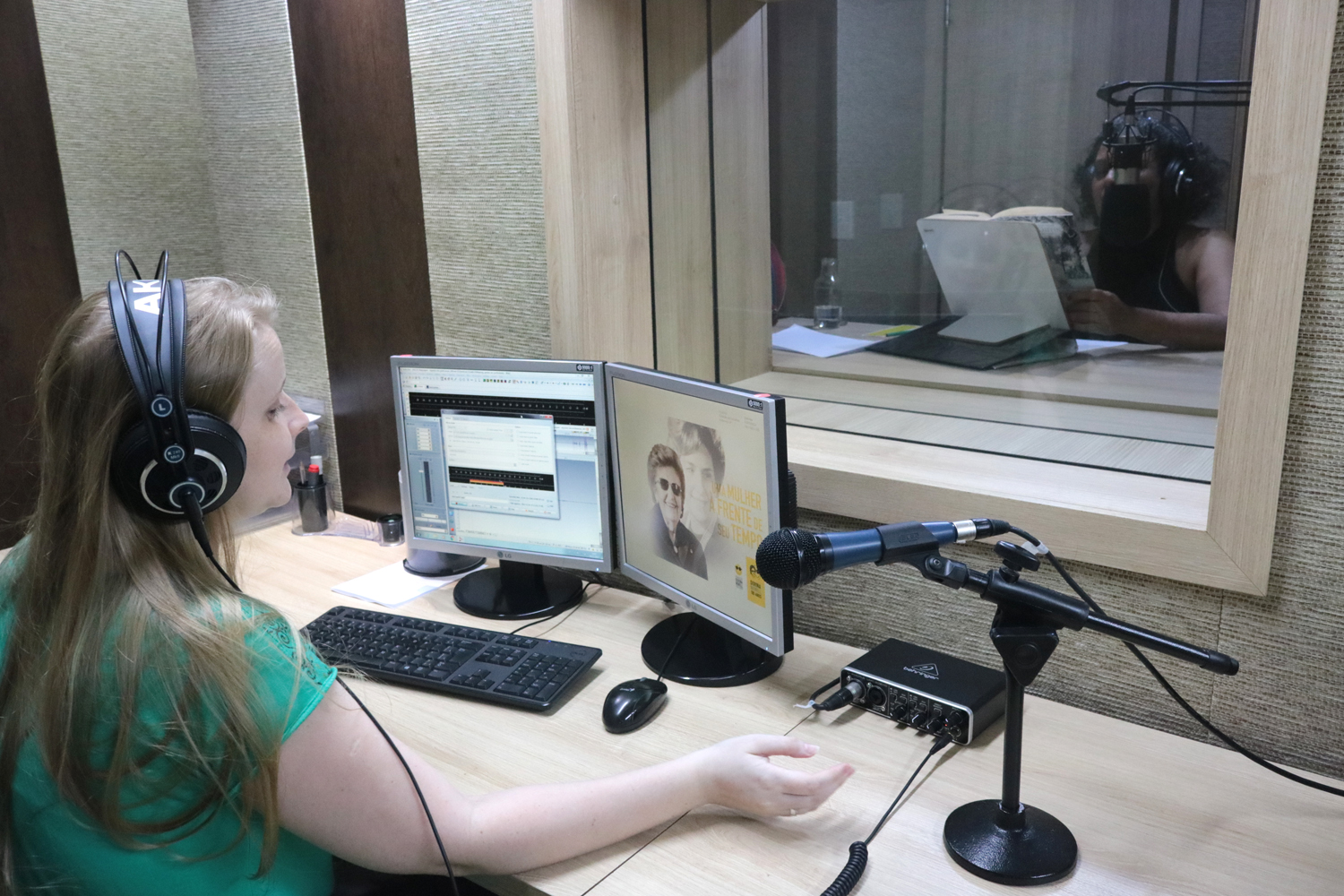 Descrição da imagem: foto de Cláudia Scheer em uma estação de trabalho com computador e microfone. Ela usa fones de ouvido. À sua frente, por uma vidraça, é possível ver a ledora Walkiria Brit dentro do estúdio.