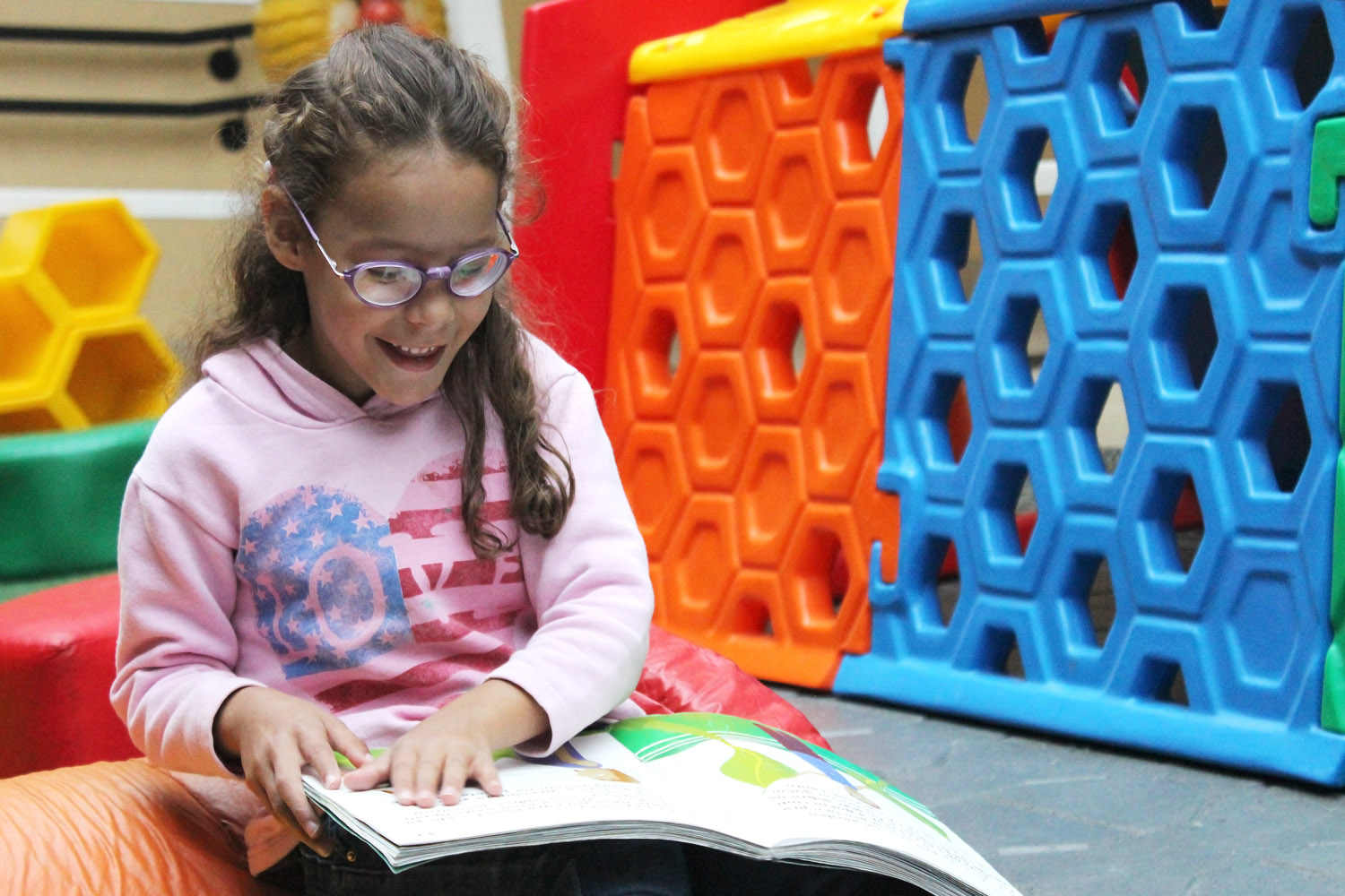 Descrição da imagem: foto de uma garotinha lendo um livro sobre as pernas. Ao fundo há brinquedos coloridos do parquinho da Fundação Dorina.