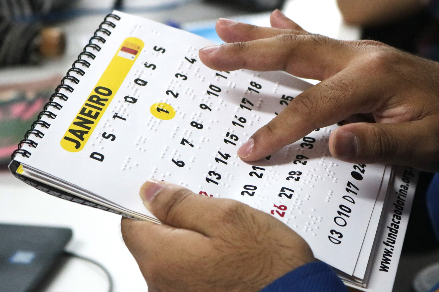 Descrição de imagem: Duas mãos segurando e tocando um calendário em braille no mês de janeiro.