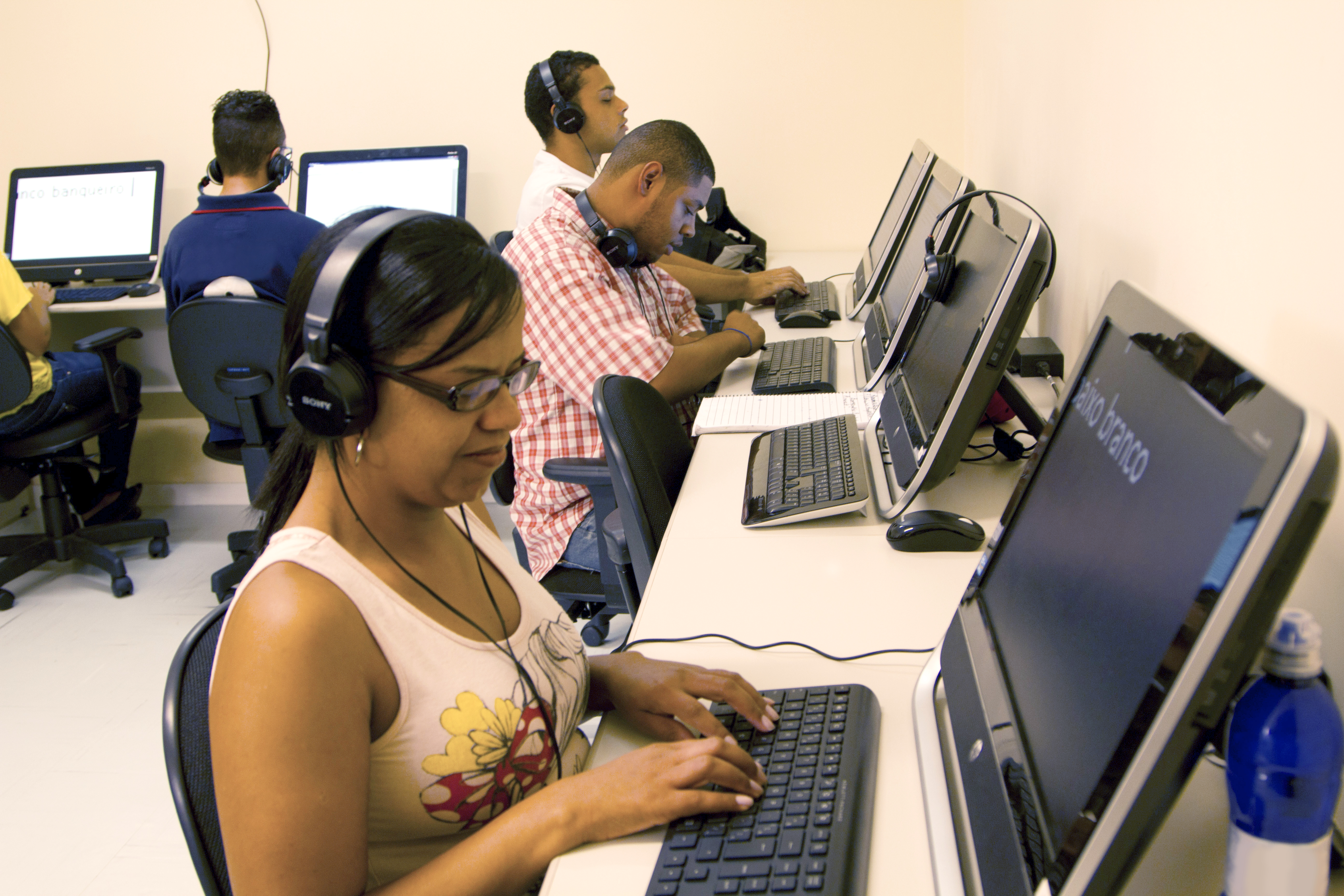 Imagem de cinco alunos da Fundação na sala de informática. Três deles, dois rapazes e uma moça, estão sentados de perfil, voltados para o lado direito. Eles utilizam headfones e estão com as mãos sob os teclados dos computadores. Outro rapaz está sentado de costas, na parede do fundo da imagem, também com headfone. Ao lado dele, está outro aluno, porém só aparece o seu braço na imagem.