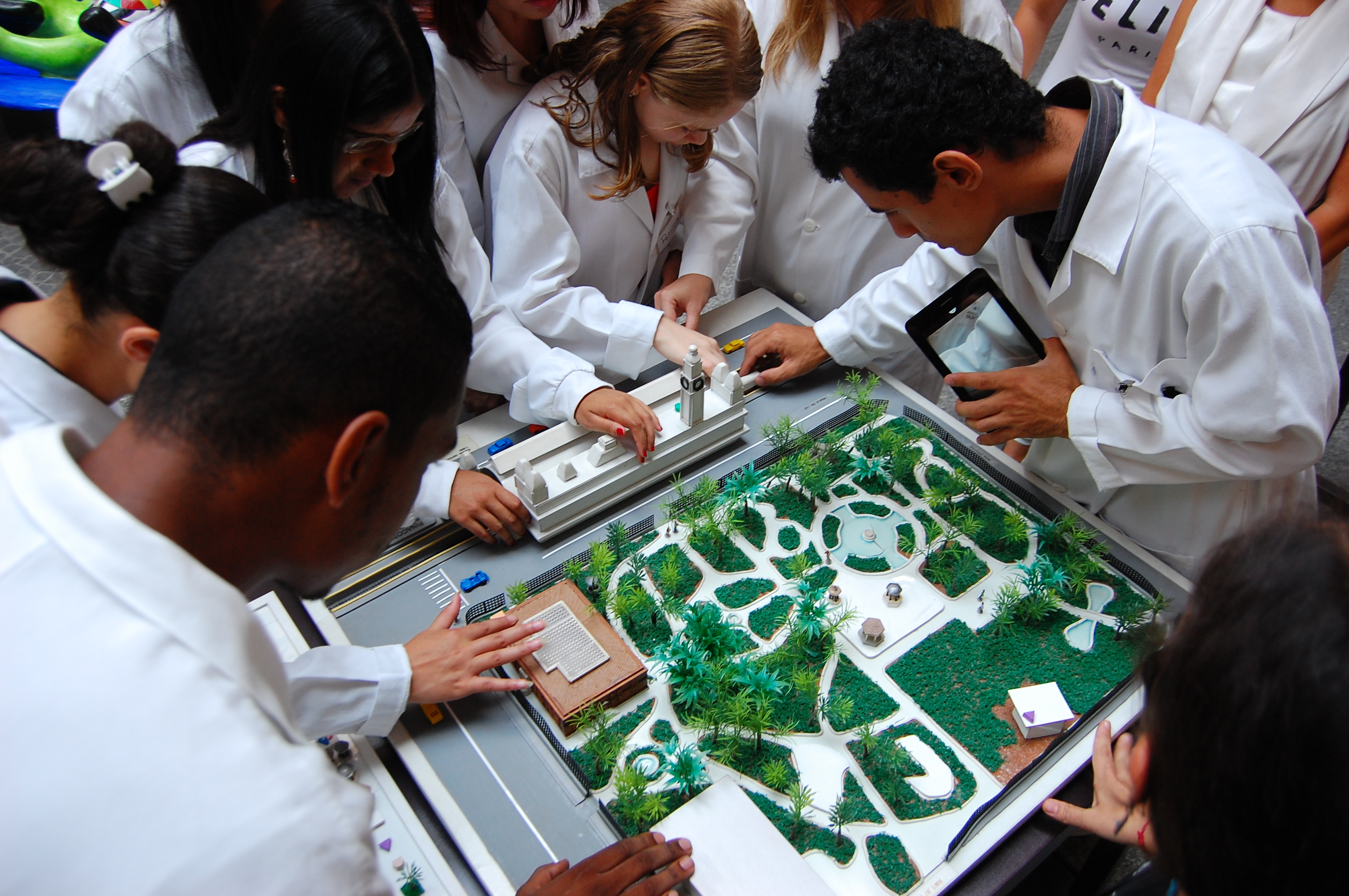 Imagem vista do alto de alunos da Fundação Dorina tateando uma maquete tátil do jardim da Pinacoteca. Eles estão de jalecos brancos, ao redor da maquete.