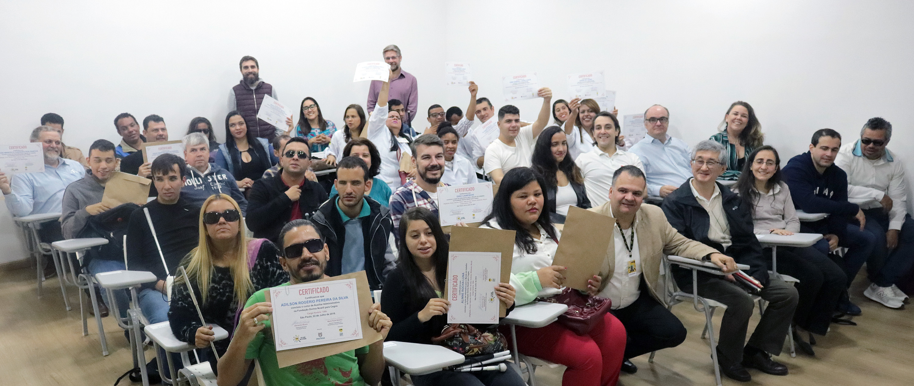 Diversos alunos no auditório da Fundação. Eles estão sentados em cadeiras, olhando para frente com seus certificados dos cursos nas mãos, alguns erguendo-os e outros mostrando-os de frente. 