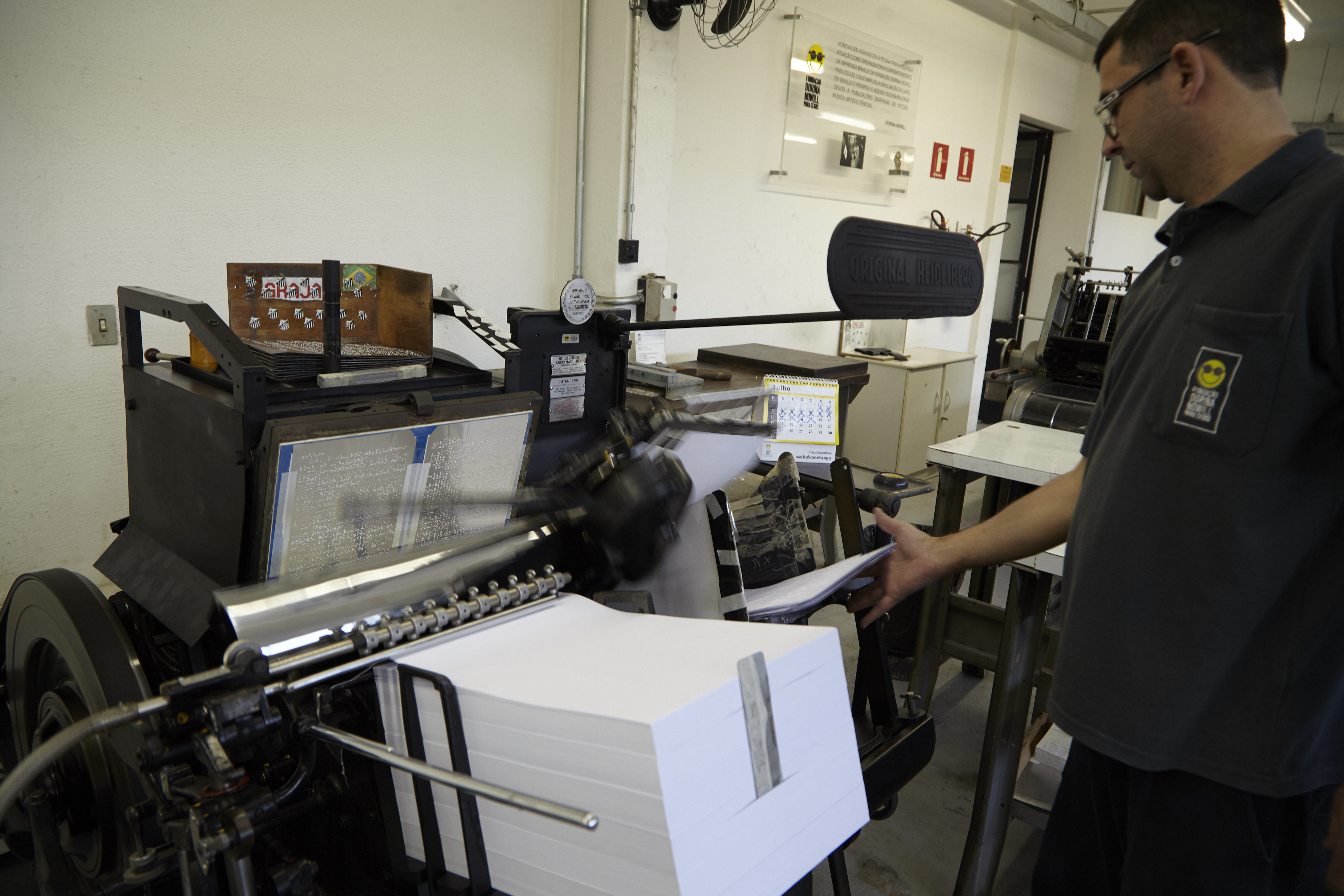 Foto de Ivo, operador da gráfica da Fundação, manuseando uma máquina de impressão em braille. Ele está no canto direito da imagem, vestindo uma camisa preta com o logotipo da Fundação. Ele está de perfil e sua mão direita está esticada até encostar em folhas de papel que estão apoiadas na máquina, que está à sua frente com uma grande pilha de papéis brancos. 