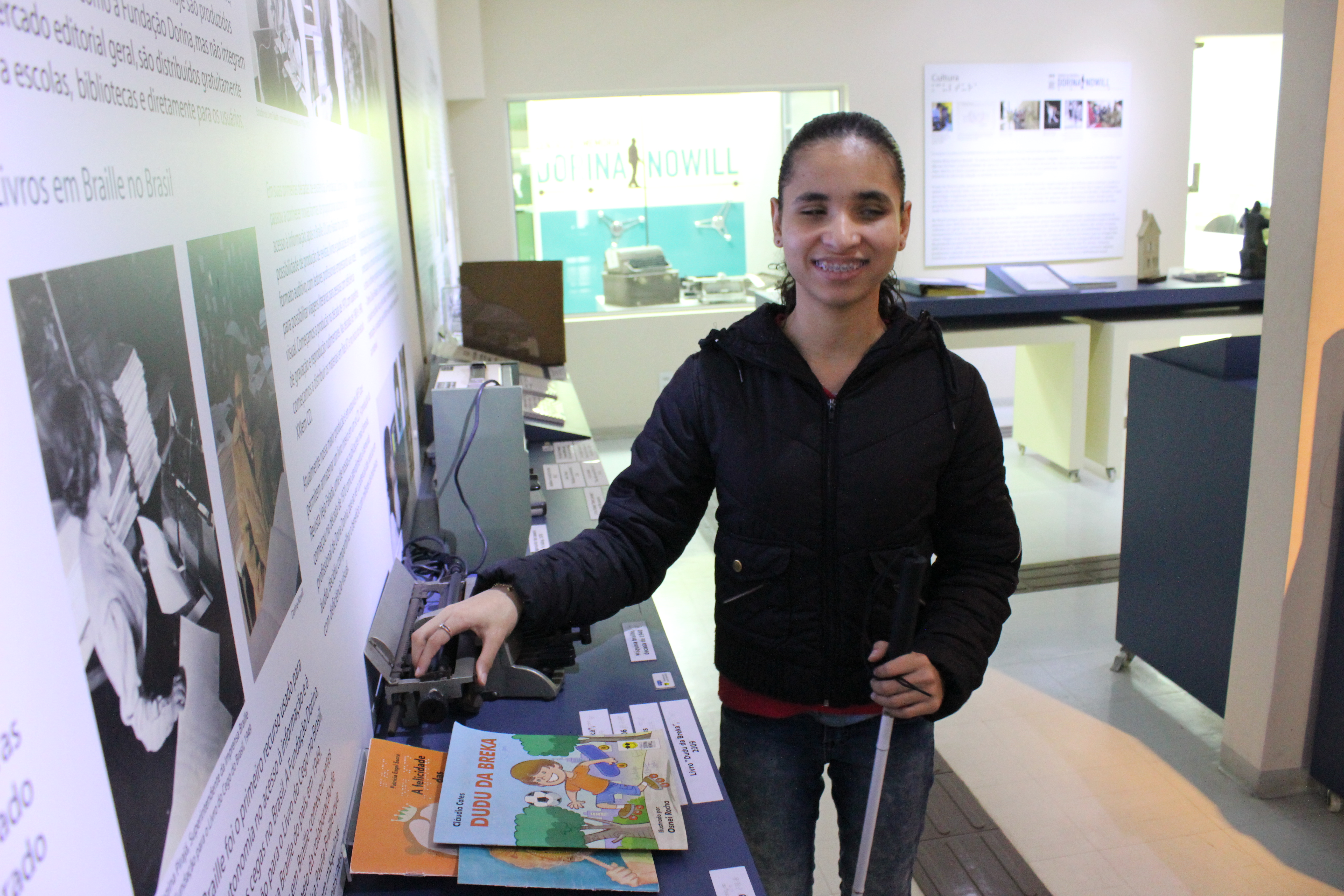 Foto de uma jovem cega na exposição do Centro de Memória. Ela veste blusa de frio preta e calça jeans preta e está com o cabelo amarrado para trás. Ela está de frente, sorrindo, segurando uma bengala branca na mão esquerda e com a mão direita ela tateia uma máquina braille antiga. A máquina está sob uma prateleira azul, ondem contém diversos livros tinta braille e outros equipamentos históricos. Na parede ao lado esquerdo da imagem, estão painéis brancos com imagens e textos da exposição. Ao fundo, aparece mais prateleiras e painéis, além de uma janela de vidro transparecendo uma estante verde. 