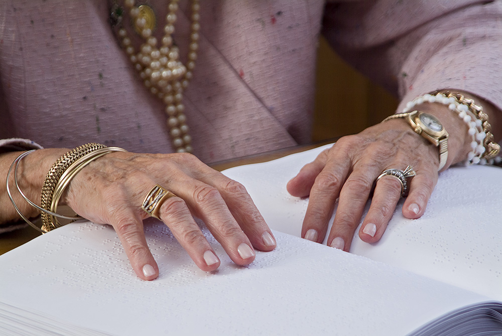 Foto das mãos de Dona Dorina sobre um livro braille. Suas mãos estão adereçadas com anéis, diversas pulseiras e um relógio. Ela veste um casaco rosa, com colar de pérolas.