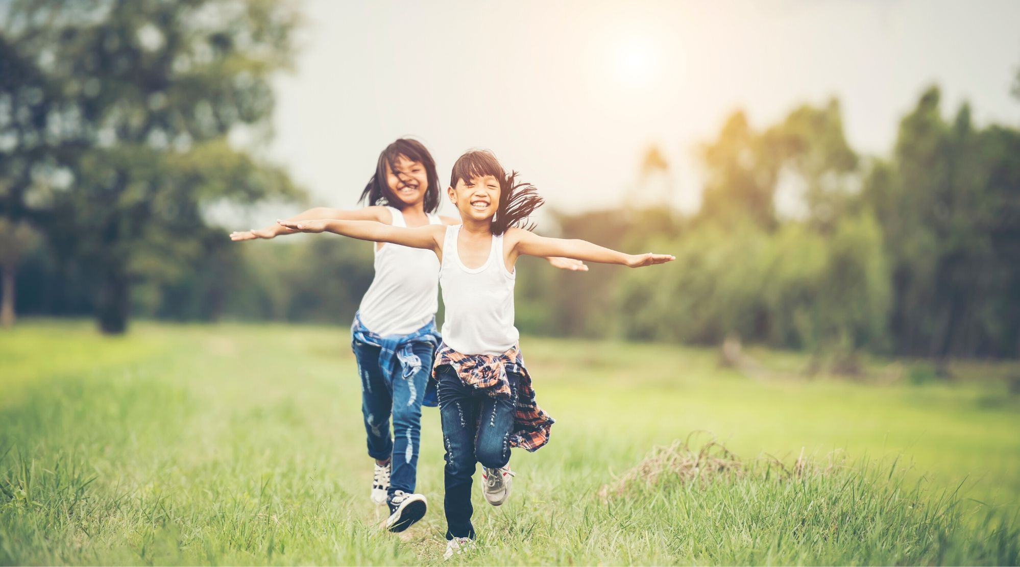 Imagem de duas crianças correndo ao ar livre. Elas estão sorrindo, de braços abertos ao redor de um campo com árvores ao fundo. Ambas as meninas têm traços asiáticos, vestem regata branca, calça jeans e blusa xadrez amarrada na cintura.