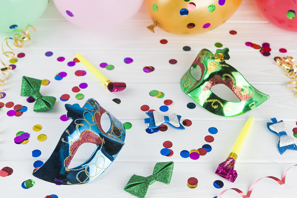 Imagem virtual de confetes e serpentinas coloridos, máscaras de carnaval e bexigas sob uma mesa branca.