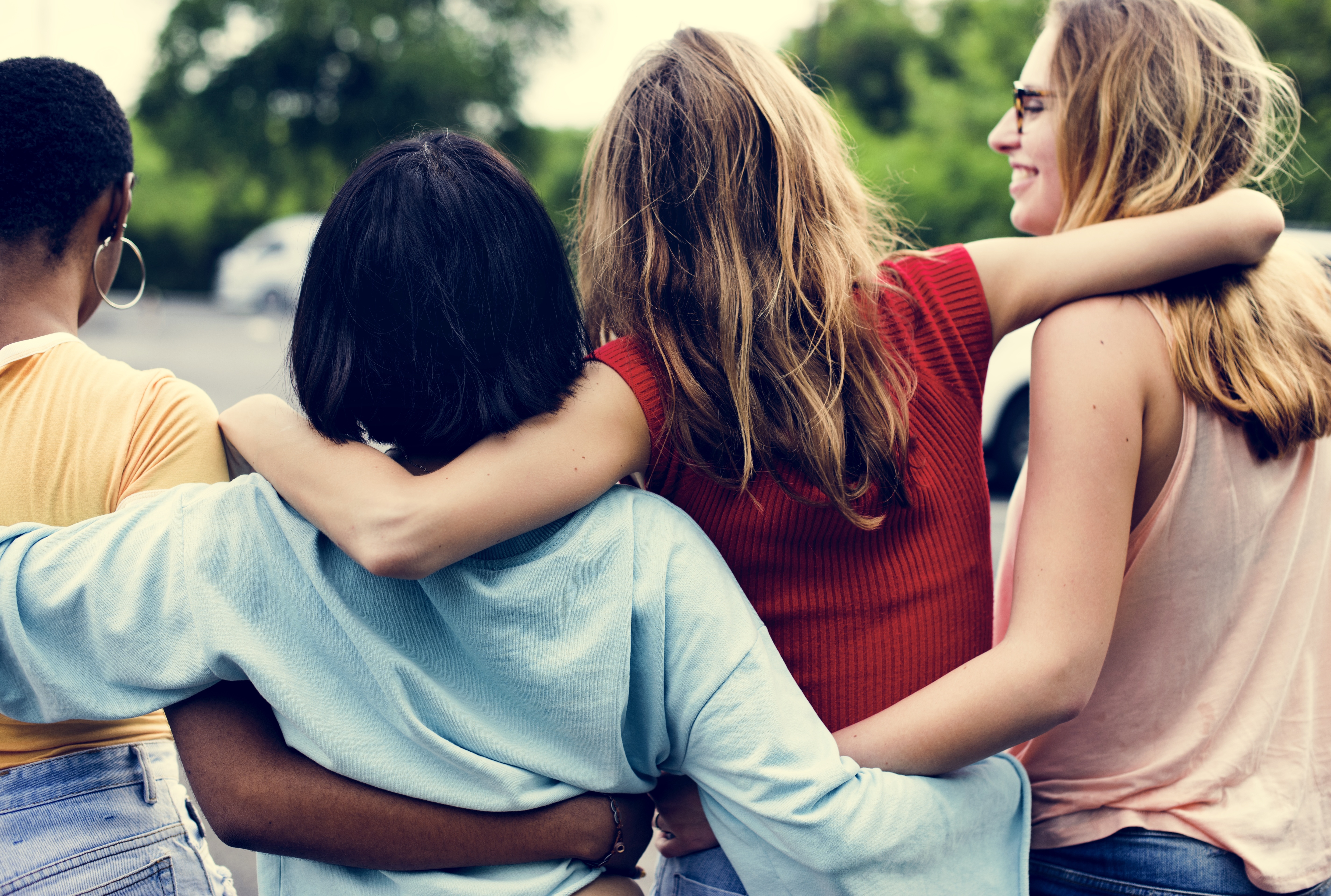 Foto de quatro mulheres andando por um parque. Ao fundo aparecem algumas árvores. Elas estão de costas, perfiladas se abraçando e uma delas está com o rosto de perfil, sorrindo..