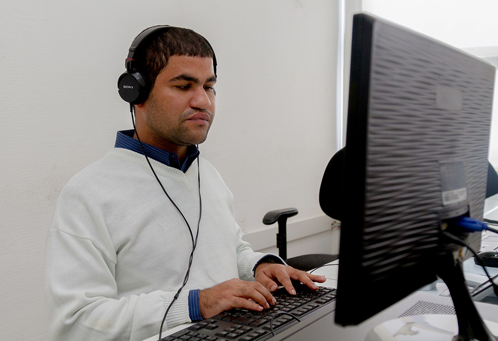 Descrição da imagem: foto de homem à frente de um computador, de olhos fechados, com as mãos sobre o teclado. Ele usa fone de ouvidos na cor preta.