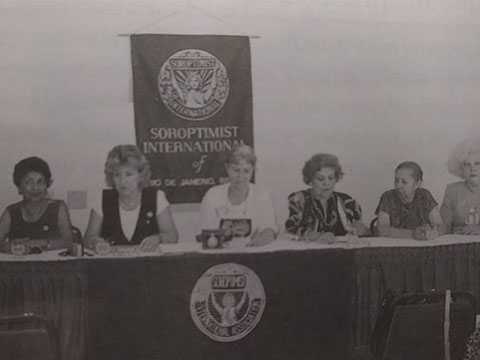 Foto antiga em preto e branco de uma reunião das soroptimistas. Há seis mulheres sentadas lado a lado apoiadas sob uma bancada. Ao fundo, há um banner com o logotipo do Soroptmist Internacional.