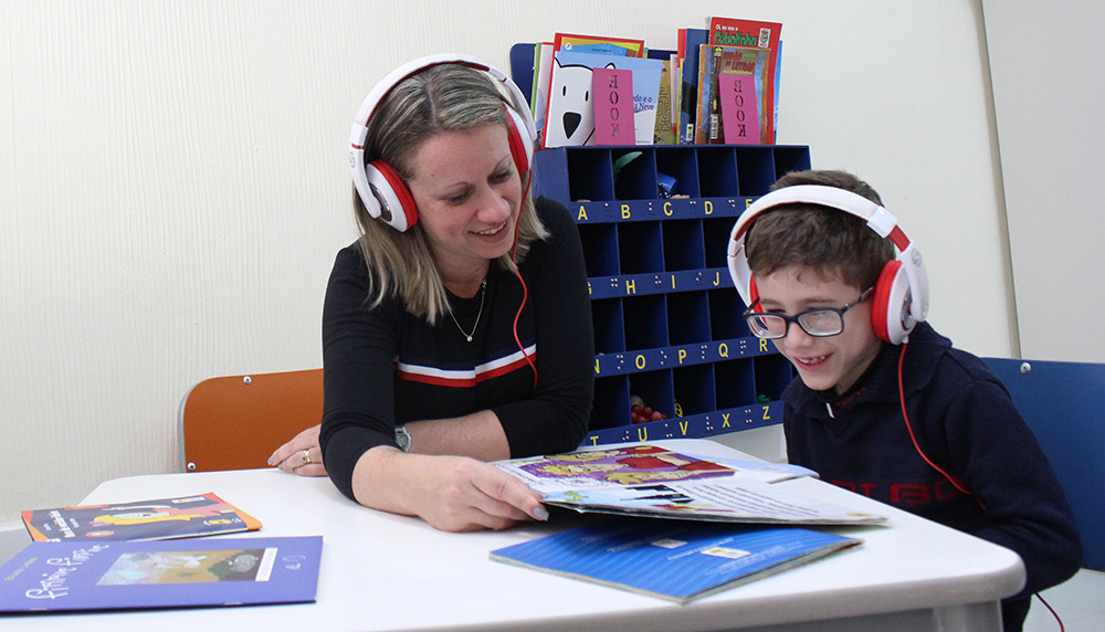 Descrição da imagem: foto de mulher e menino com livros abertos sobre a mesa. Eles usam fones de ouvido. O garoto usa óculos. Eles estão olhando para o livro e sorrindo.