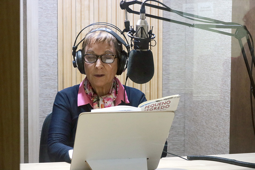 Descrição da imagem: foto de Heloísa Schurmann com um livro aberto sobre prancheta à sua frente. Ela usa fones de ouvido e tem um microfone de estúdio posicionado à altura do rosto.