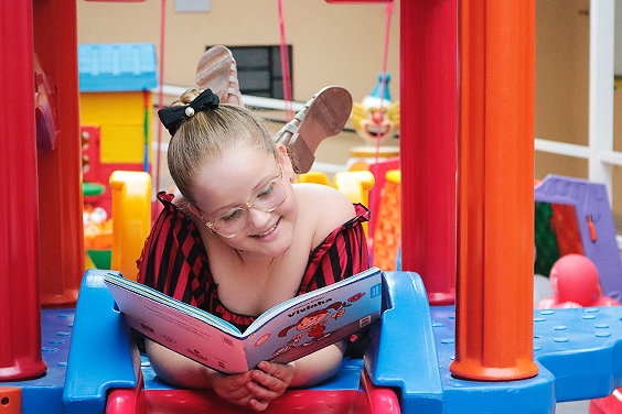 Descrição da Imagem: foto de Isabella, criança atendida pela Fundação, no parquinho. Ela está deitada, com as pernas cruzadas para o alto e com um livro infantil aberto à sua frente. Ela sorri ao ler o livro.