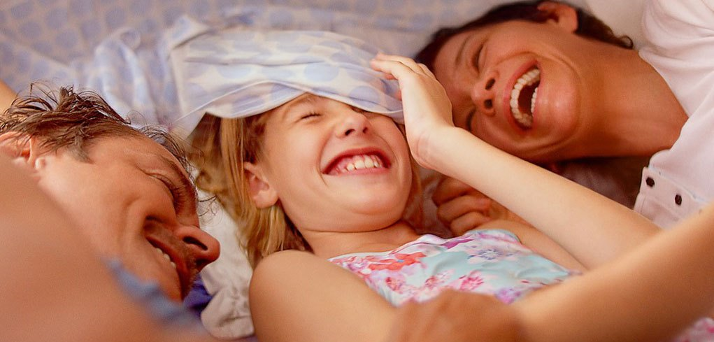 Cena do filme Pequeno Segredo. Um casal está deitado na cama com sua filha no meio, ambos aparecem do busto para cima e todos estão sorrindo.