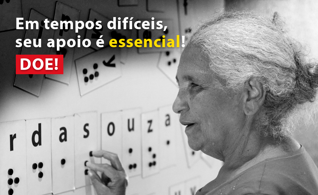 Descrição da imagem: foto em preto e branco de uma senhora de cabelos brancos de perfil, tocando um painel na parede com o alfabeto em braille. Ao lado esquerdo há o texto "Em tempos difíceis, seu apoio é essencial. Doe!"