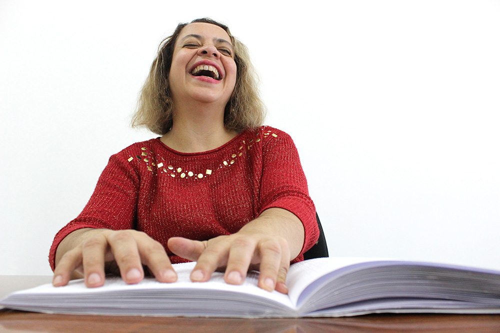 Descrição da imagem: foto de mulher tateando um livro em braille aberto sobre a mesa e sorrindo.