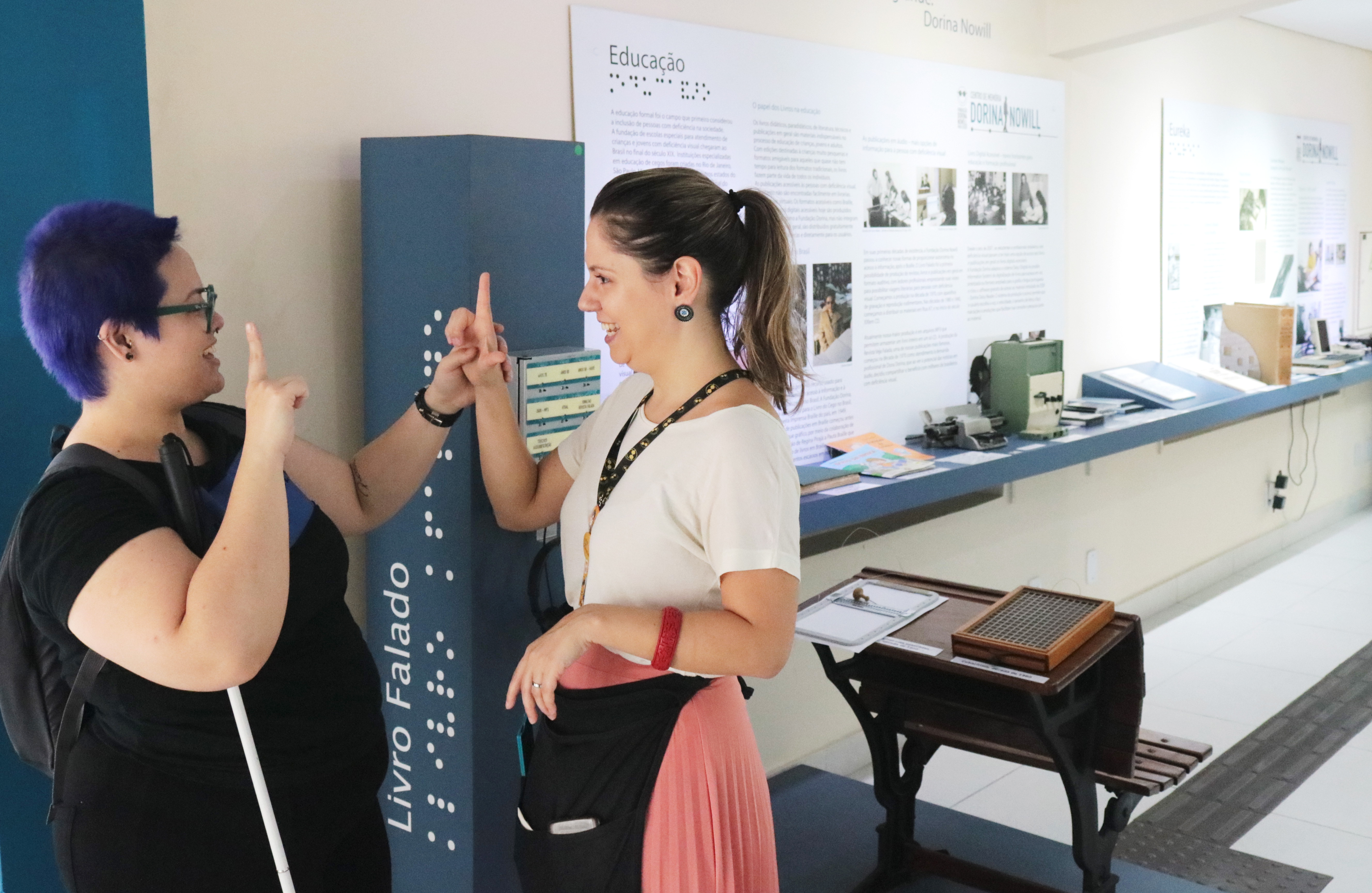 Foto de atendimento no Centro de Memória Dorina Nowill. Silvia, educadora do Centro de Memória, está conversando em libras com uma visitante e, ao fundo, aparecem algumas peças da exposição. 