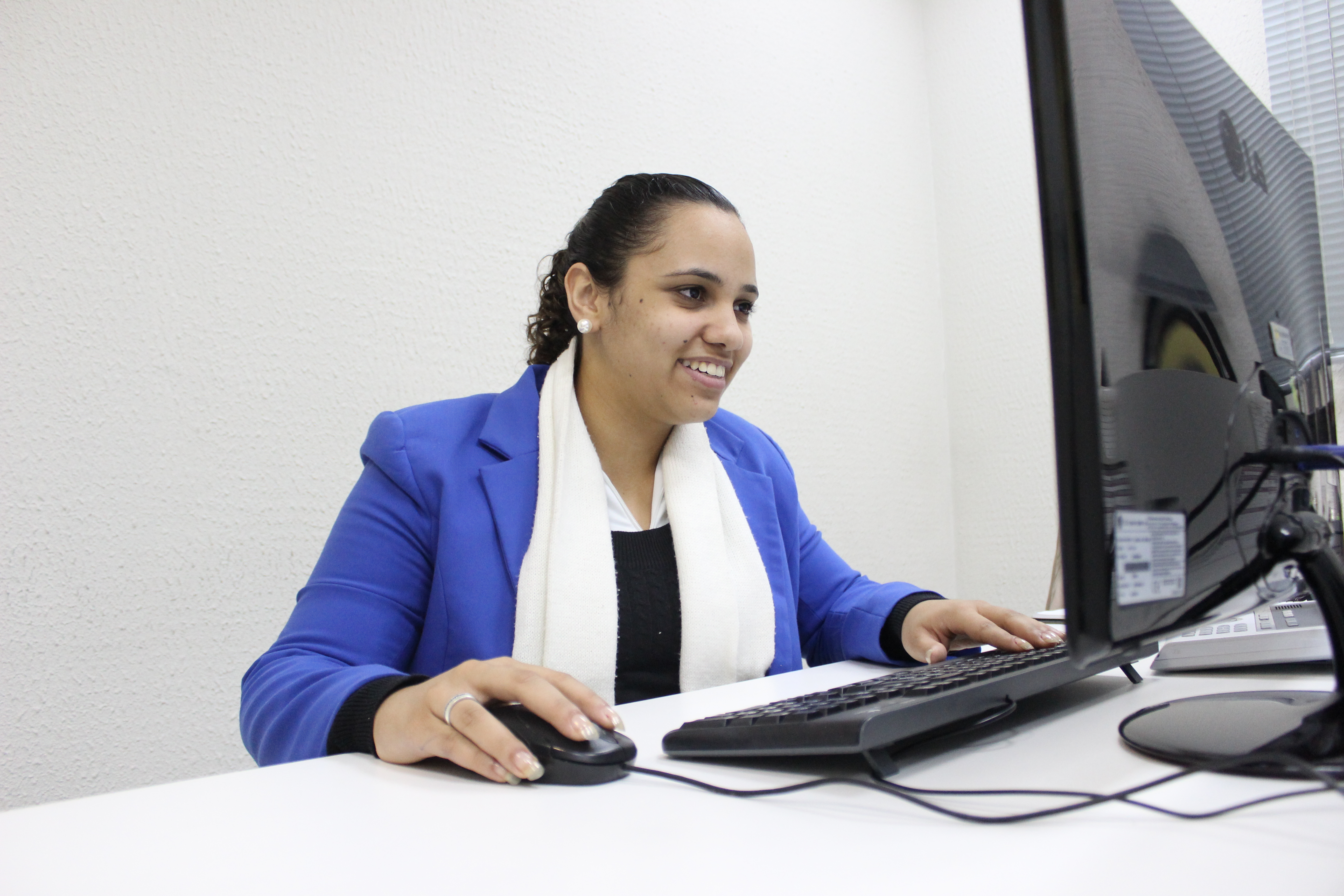 Foto de Stephanie, cliente atendida pela Fundação. Ela está sentada em frente a uma mesa branca, com uma tela de computador a sua frente. Suas mãos estão sb um teclado e um mouse pretos. Stephanie veste bleaser azul e cachecol branco e sorri.