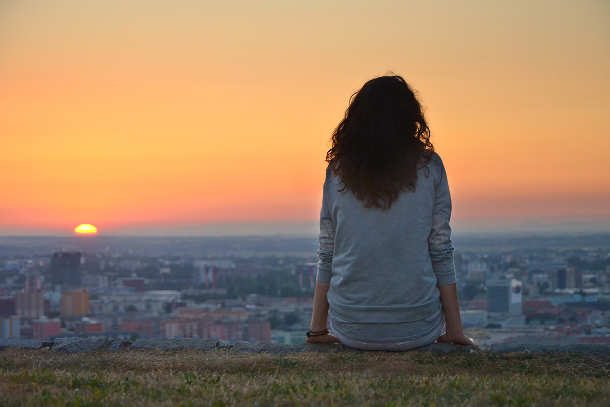 Descrição da imagem: foto de mulher de costas sentada numa mureta observando o por do sol. Ela está em um ponto alto de uma cidade, com uma vista repleta de casas e prédios lá em baixo.