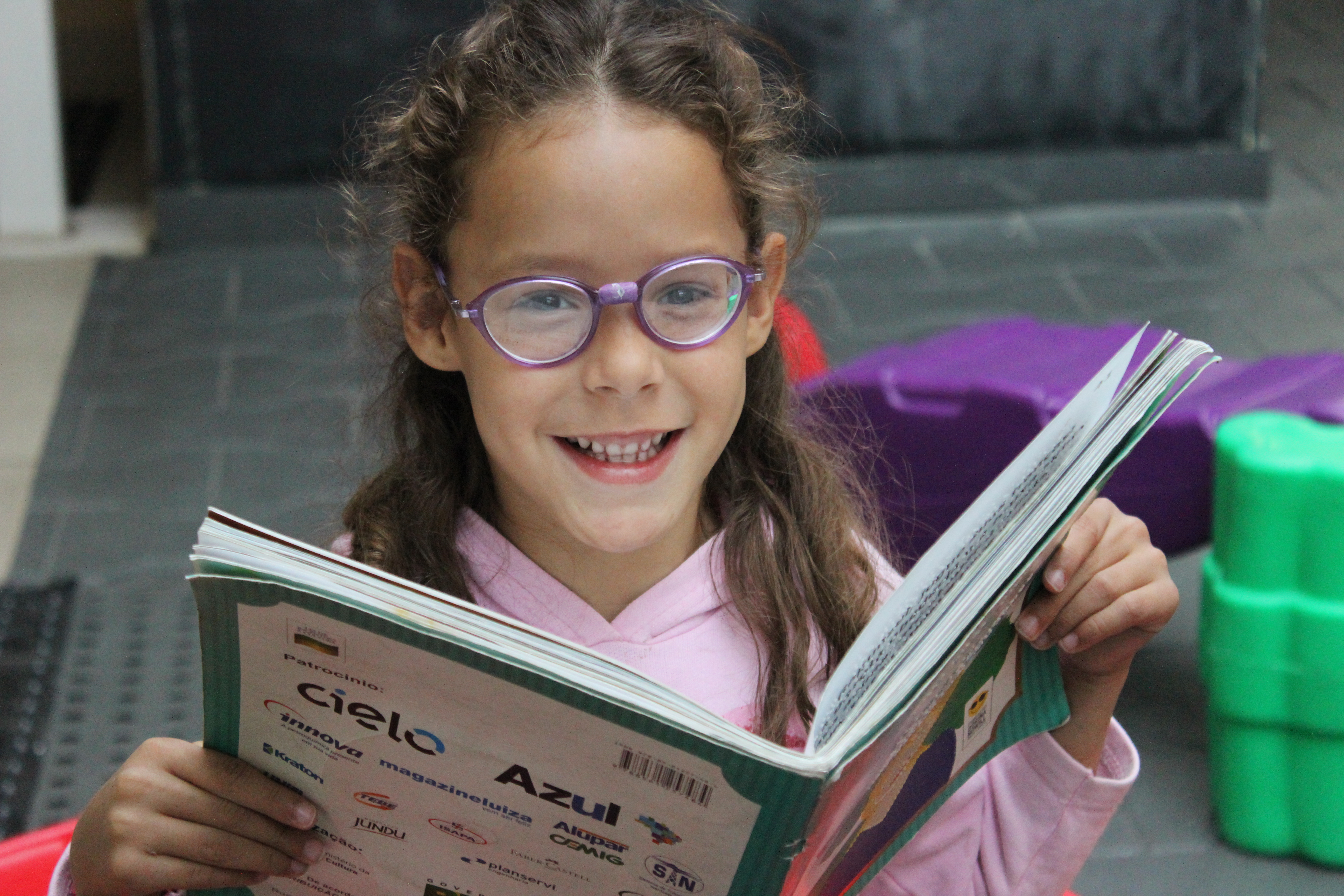 Foto de Ana Júlia, criança atendida pela Fundação. Ela está no parquinho da Fundação, segurando um livro aberto em sua frente e sorri. Ela veste blusa rosa e óculos de hastes na cor roxa.