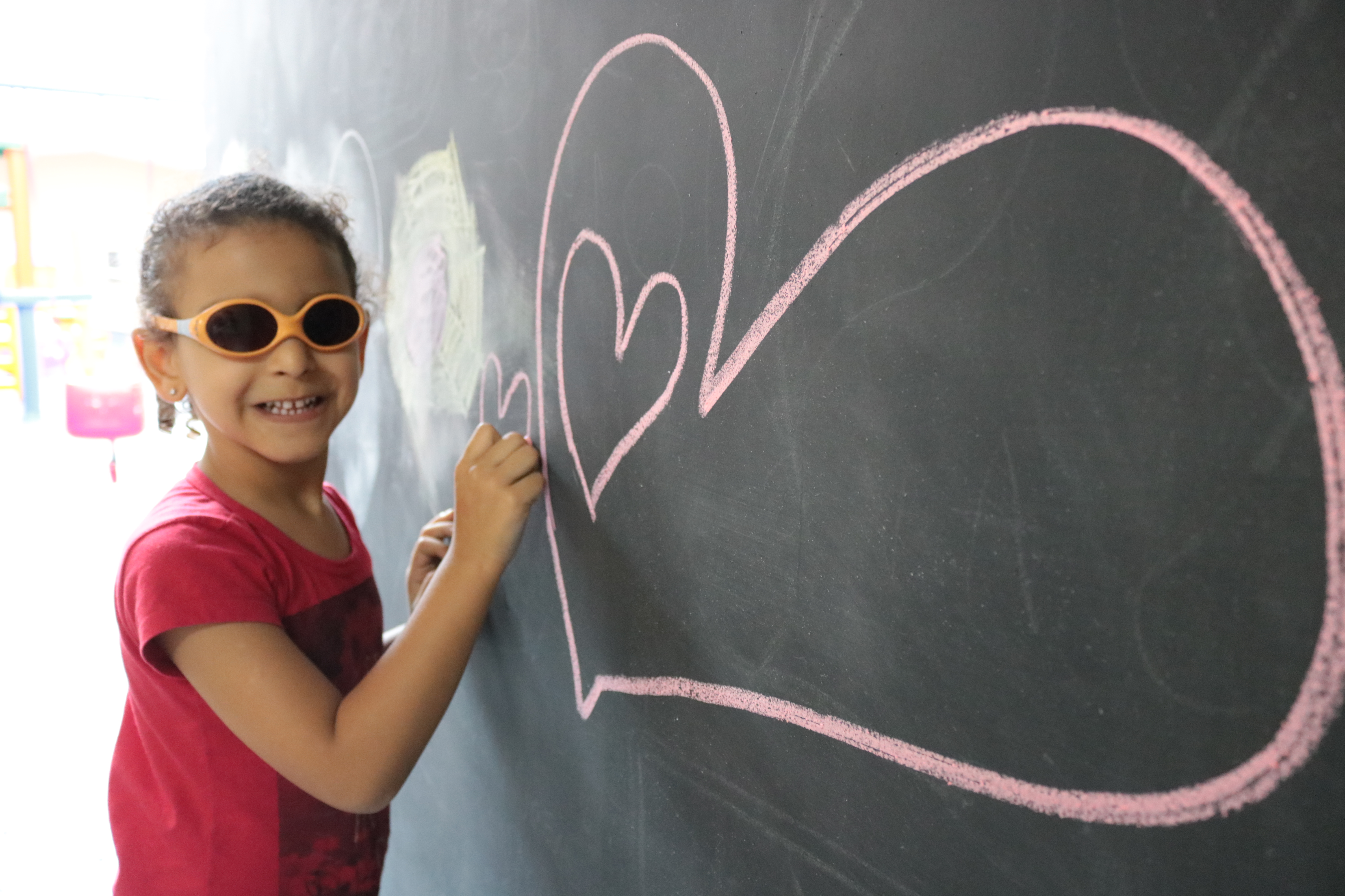 Descrição da imagem: foto de menina sorridente de óculos escuros. Ela segura um pedaço de giz sobre um quadro negro onde está desenhado um coração na cor rosa.