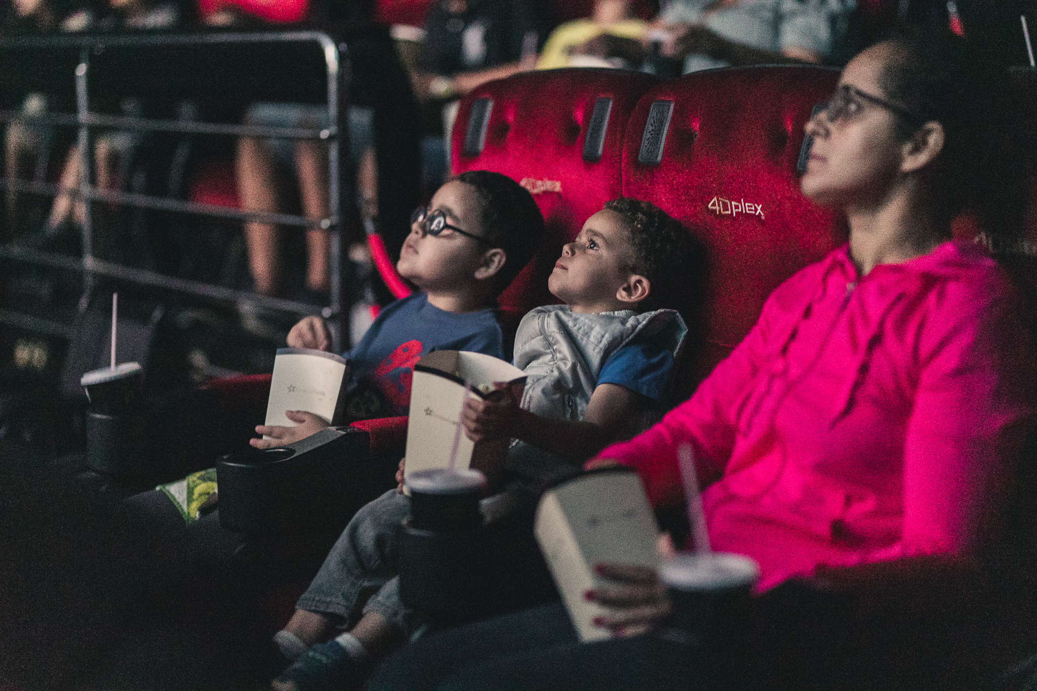 Imagem de uma sessão de cinema inclusivo promovido pelo Telecine. Em destaque, há dois meninos e uma mulher sentados em poltronas de cinema. Eles olham para frente e estão segurando embalagens de pipoca nas mãos.