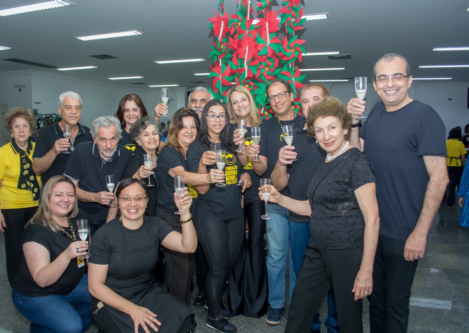 Imagem de diversas pessoas, entre homens e mulheres voluntários e colaboradores, no Bazar da Costura 2019. Eles vestem calças e camisetas pretas, a maioria com o logotipo da Fundação Dorina. Eles estão reunidos em pé, sendo que duas mulheres estão à frente agachadas. Todos estão segurando um taça de champanhe nas mãos.