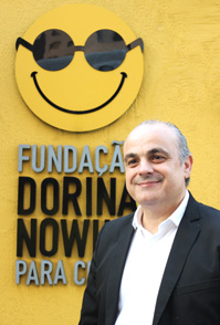 Foto de Marcelo Panico, advocacy da Fundação Dorina. Ele está representado do tórax para cima, veste terno, camisa branca e sorri. Ao Fundo, está uma parede amarela com o logotipo da Fundação Dorina em relevo. 