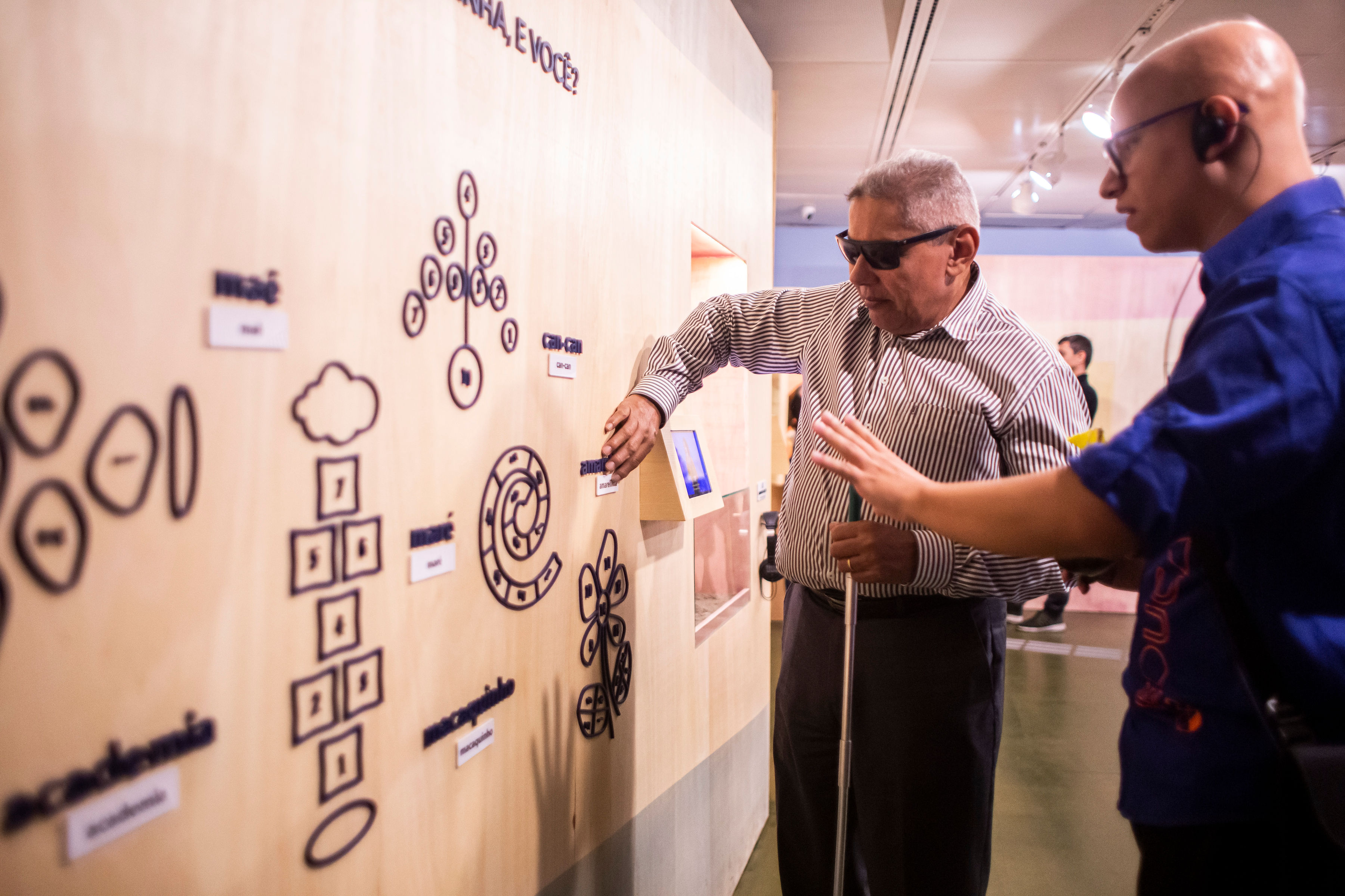 Imagem de uma exposição no Itaú Cultural. A direita há uma parede com desenhos em relevo e à esquerda um homem tateando a parede com a mão direita. Ele veste camisa social listrada de manga longa e calça preta, usa óculos escuros e segura uma bengala na mão esquerda. Ao seu lado, está outro homem com o braço esticado para frente. Ele veste camisa azul, calça preta e utiliza um aparelho de comunicação no ouvido.