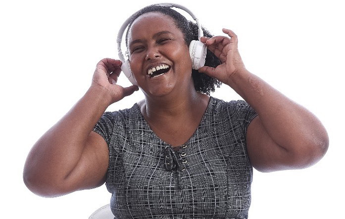 Descrição da imagem: foto de Fernanda, uma mulher negra retratada da cintura pra cima, com fones de ouvido brancos na cabeça. Ela toca o equipamento com as duas mãos e exibe um largo sorriso de olhos fechados.